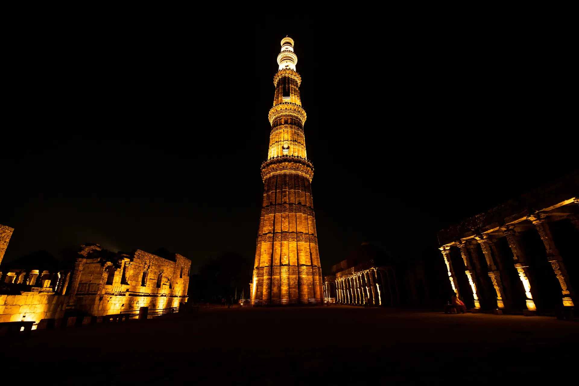 Qutub Minar illuminated in gold against the night sky, flanked by the lit ruins of the Qutub complex in New Delhi