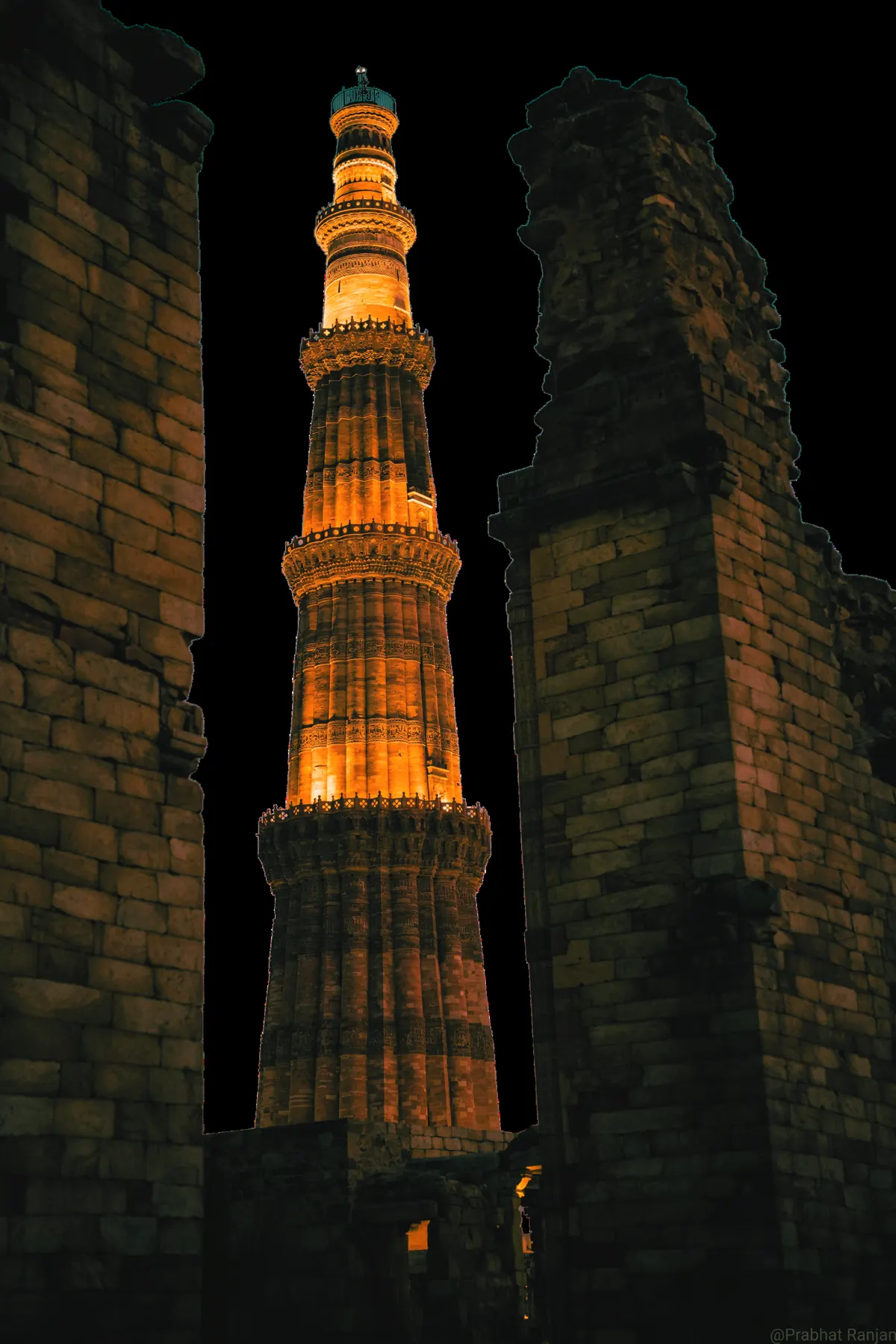 The Qutub Minar lit in gold, framed between dark stone ruins at night